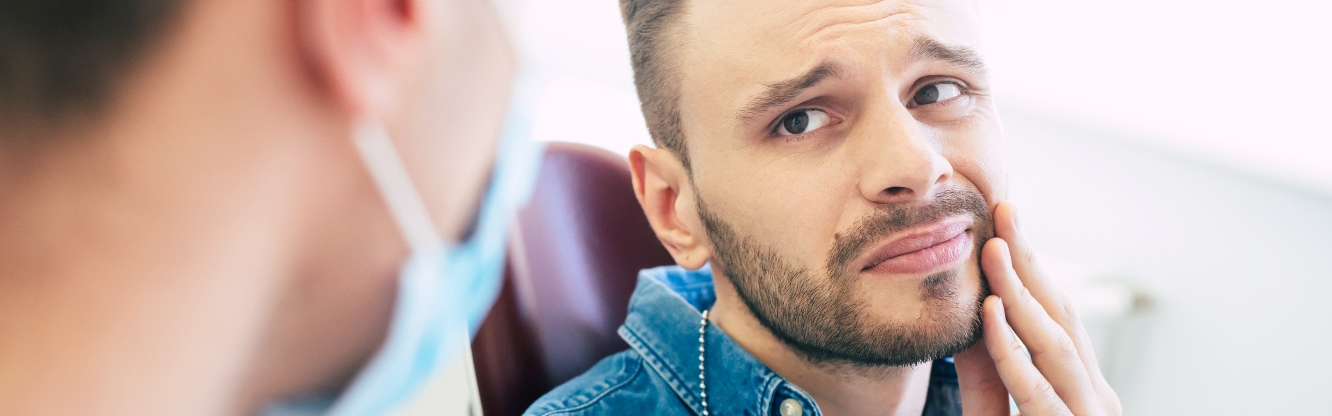 Symptoms of gums pain a man with a worried face is holding his hand on his cheek because of irritating pain in front of a dentist who is going to give a patient a treatment jpg